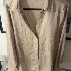 Beige skjorta från H&M med knappar - Stilren beige skjorta från H&M i storlek S. Skjortan har klassisk krage, långa ärmar och knäppning framtill med matchande knappar. Materialet är mjukt och lätt, perfekt för en chill och clean look. Passar dig som gillar enkel och tidlös stil.