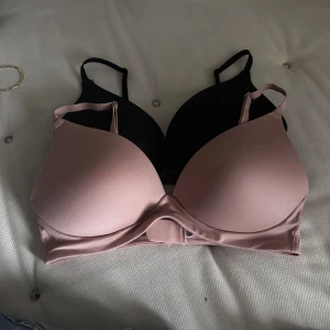 Svart och rosa bh från Primark - Två bh-ar från Primark, en i svart och en i ljusrosa. Båda är släta och har justerbara axelband samt mjuka kupor utan bygel. Materialet är lent och syntetiskt, perfekt för en clean look under kläderna. Säljes som set! Oanvända, rosa är endast testad. 