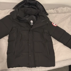Svart dunjacka från Canada Goose - Säljer en svart dunjacka från Canada Goose med klassisk patch på ärmen. Jackan har huva, dragkedja och knappar framtill samt flera fickor. Perfekt för kalla vinterdagar och riktigt snygg streetstil. Jackan är använd 1 vinter och det finns ett litet håll runt nack krogen annars är den i mycket bra skick. Kan frakta paketet eller så kan jag mötas upp i gbg eller i alingsås kon dm för mer info. tar även emot swish