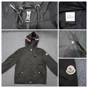 Svart vindjacka från Moncler med huva - Svart vindjacka från Moncler med huva och dragsko, snygg detalj med röd och vit rand på huvkanten. Jackan har dragkedja framtill, två fickor med tryckknappar och Moncler-logga på ärmen. Perfekt för dig som vill ha en stilren och sportig look.