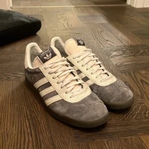 Adidas Gazelle i grå mocka och vitt - Säljer ett par Adidas Gazelle sneakers i grå mocka med vita läderdetaljer och klassiska tre ränder på sidorna. Skorna har vit plös, snörning och mörk yttersula. Perfekta för dig som gillar retrostil och vill ha något som sticker ut lite extra. Använda fåtal gånger. 