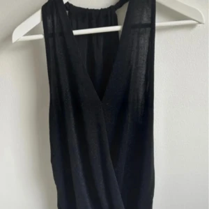 Svart glittrigt omlottlinne - Svart linne omlott med glittrigt tyg och djup v-ringning. Linnet är ärmlös och har en snygg halterneck med rynkad detalj i nacken. Perfekt för dig som vill sticka ut med en elegant och trendig look.