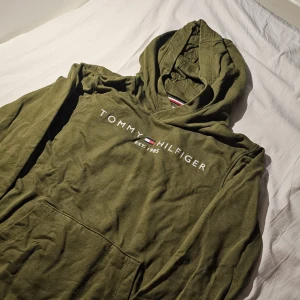 Olivgrön hoodie från Tommy Hilfiger - Snygg olivgrön hoodie från Tommy Hilfiger med klassisk logga och text på bröstet. Tröjan har huva och en stor magficka framtill. Perfekt för dig som gillar en avslappnad och trendig stil.