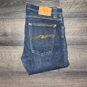 Nudie jeans  - Snygga mörkblå jeans från Nudie Jeans med klassisk femficksdesign och gul kontrastsöm på bakfickorna. Modell TUBE TOM  Jeansen är tillverkade i stretchig denim för extra komfort. Midja 39cm, längd 101cm. JAG SKICKAR EJ FLER BILDER NÄR JAG BÄR JEANSEN! för storleksguide hänvisar jag till måtten och för bättre inblick i passformen rekommenderar jag att googla på modellnamnet :) s3