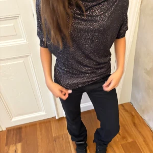 Svart glittrig topp med prickigt mönster - Svart topp med glittriga silverprickar och lös passform. Toppen har korta ärmar och en draperad, något v-ringad halsringning. Materialet är mjukt och stretchigt, perfekt för att sticka ut med en snygg look.