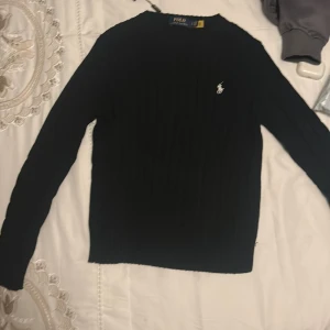 Svart stickad tröja från Polo Ralph Lauren finns i grå och Lilla - Svart stickad tröja från Polo Ralph Lauren med klassisk rund hals och vit broderad logga på bröstet. Tröjan har lång ärm och ribbade muddar, perfekt för lager-på-lager under höst och vinter. Snygg och tidlös modell som funkar till allt.