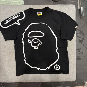 Svart BAPE t-shirt med stor ape-print - Svart t-shirt från BAPE med ikoniskt stort vitt ape-ansikte tryckt på framsidan och cool textbubbla på axeln. Klassisk passform, korta ärmar och crewneck. Tillverkad i mjuk bomull och har BAPE-logga på ärmen. Storlek är L men passar M