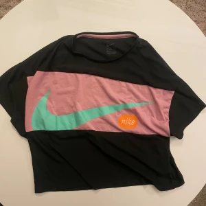 Svart och rosa croppad topp från Nike - Cool croppad topp från Nike i svart och rosa med stor turkos swoosh-logga och orange Nike-detalj på framsidan. T-shirten har korta ärmar och är gjord i mjuk bomull. Perfekt för dig som gillar sportig streetstyle och vill sticka ut med färg.