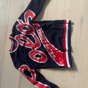 Mörkblå tröja från Ralph Lauren - Snygg mörkblå tröja från Ralph Lauren med stora röda och vita tryckdetaljer och logga framtill. Tröjan har långa ärmar och ribbade muddar i rött, vitt och blått. Perfekt för dig som gillar sportig och trendig stil.