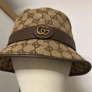 Beige bucket hat från Gucci - Säljer en beige bucket hat från Gucci med klassiskt GG-monogram i brunt. Hatten har ett brunt läderband runtom med GG-logga i metall framtill. Tillverkad i bomull och läder, perfekt för dig som vill sticka ut med en lyxig accessoar.