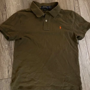 Olivgrön pikétröja Polo Ralph Lauren - Snygg olivgrön pikétröja från Polo Ralph Lauren i slim fit-modell. Klassisk krage, korta ärmar och knäppning framtill. Ikoniska broderade loggan i orange på bröstet. Tillverkad i mjuk bomull för en skön känsla. Passar dig som är runt 160.