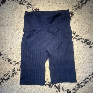 Mörkblå ribbade shorts från Stronger - Säljer ett par mörkblå, ribbade cykelshorts från Stronger med diskret logga i midjebandet. Shortsen är stretchiga och perfekta för träning eller chill. Materialet är mjukt och följsamt syntet, med bred midjeresår för extra komfort.