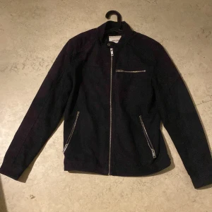 Svart jacka från Jack & Jones - Stilren svart jacka från Jack & Jones med raka linjer och tre dragkedjefickor framtill. Jackan har hög krage och hel dragkedja, perfekt för dig som gillar en clean look. Materialet känns slitstarkt och passar till många outfits.