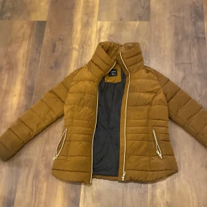 Brun pufferjacka från Lindex - Säljer en brun pufferjacka från Uniqlo med guldfärgade dragkedjor och hög krage. Jackan är quiltad och har två fickor med dragkedja. Perfekt för kyliga dagar och har en snygg, enkel design.