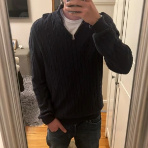 Mörkblå stickad tröja John Henric - Säljer en mörkblå stickad tröja från John Henric med kabelstickat mönster och half zip-dragkedja vid halsen. Tröjan har lång ärm och ribbade muddar, perfekt för lager-på-lager och snygg till jeans.