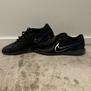 Svarta Nike fotbollsskor med blå detalj - Säljer ett par svarta Nike fotbollsskor med silverfärgad swoosh och blå detalj på sidan. Skorna har snörning och är tillverkade i syntetmaterial med mönstrad sula för bra grepp på planen. Perfekta för dig som vill ha stilrena och sportiga skor till fotbollsträningen. Dom har en skavank på sista bilden som ni ser.