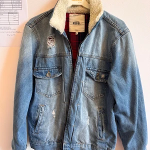 Ljusblå jeansjacka med teddykrage Rebel - Snygg ljusblå jeansjacka från Redefined Rebel med vit teddykrage och röd-rutigt foder. Jackan har slitna detaljer, två bröstfickor med knappar och dragkedja framtill. Perfekt för dig som gillar en avslappnad och trendig stil.