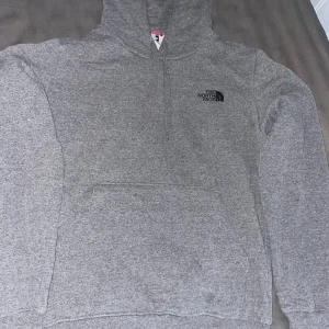 Grå hoodie från The North Face - Säljer en grå hoodie från The North Face i storlek S. Tröjan har huva med snörning, stor magficka och mjukt fleecefoder på insidan. Diskret logga på bröstet och klassisk passform. Perfekt för chill dagar eller när du vill vara extra bekväm.