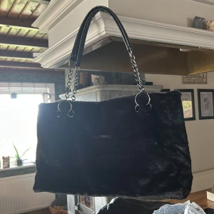 Svart pälsig axelväska med kedjehandtag - Stor svart axelväska i mjuk fuskpäls med rejäla kedjehandtag och svarta läderdetaljer. Väskan har en rektangulär form och ger en lyxig vibe, perfekt för dig som vill sticka ut. Enkel design utan synliga loggor eller mönster.