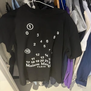 Maison Margiela svart t-shirt med print - Svart t-shirt från Maison Margiela med ikoniskt siffror-print och logga på framsidan. Klassisk rund hals och korta ärmar. Tillverkad i mjuk bomull, med de typiska vita stygn på ryggen. Perfekt för dig som gillar statement-plagg och designer vibes.