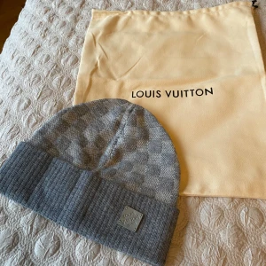 Grå Louis Vuitton mössa i ull - Exklusiv grå mössa från Louis Vuitton med klassiskt rutigt mönster och ribbad uppvikt kant. Mössan har en metallbricka med Louis Vuitton-logga framtill och är tillverkad i mjuk ull. Perfekt accessoar för att lyfta din vinterstil.