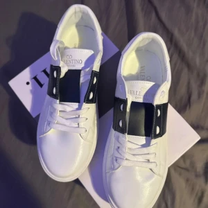 Valentino Garavani vita sneakers - Snygga vita sneakers från Valentino Garavani med bred mörkblå rem över ovansidan och coola nitar längs sulan. Skorna har rund tå, platt sula och snörning. Tillverkade i skinn för en lyxig känsla och stilren look. Helt nya aldrig använda. Storlek 41-42. Pris kan diskuteras.