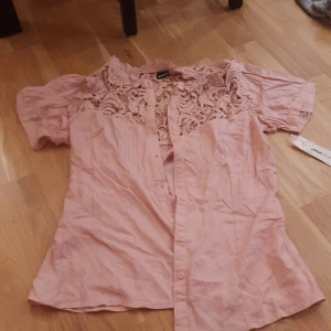 Rosa spetsblus från Gina Tricot - Säljer en ljusrosa blus från Gina Tricot med korta puffärmar och knappar framtill. Överdelen har snygga spetsdetaljer och blusen är tillverkad i mjuk bomull. Perfekt för dig som gillar romantisk och feminin stil.