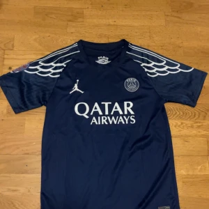 PSG x Jordan blå fotbollströja - Snygg mörkblå PSG fotbollströja i samarbete med Jordan. Tröjan har korta ärmar med vitt vingmönster på axlarna, Paris Saint-Germain-logga på bröstet och Jordan-logga. Materialet är lätt och andas, perfekt för träning eller match.