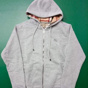 Grå hoodie från Burberry med dragkedja - Snygg grå hoodie från Burberry i storlek M med klassisk rutmönstrad insida vid huvan. Tröjan har dragkedja framtill, snörning vid huvan och två fickor. Perfekt för dig som gillar stilrena och exklusiva märken.