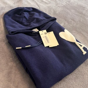 Ami Paris tröja  - Säljer en mörkblå hoodie från Ami Paris med ikoniskt vitt hjärta och A på bröstet. Tröjan har huva med snörning, känguruficka och är stickad i mjukt material. Perfekt för dig som vill ha en stilren och trendig look.