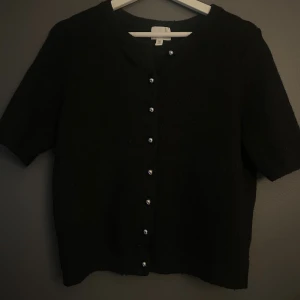Svart kofta med kort ärm och knappar - Svart kofta från H&M med korta ärmar och rund hals. Tröjan har snygga silverfärgade knappar framtill och är stickad i mjukt material. Knappt använd💛