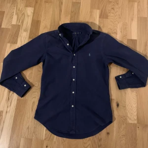 Mörkblå skjorta från Ralph Lauren - Mörkblå skjorta från Ralph Lauren i slim fit med klassisk button-down krage och vita knappar. Skjortan är i storlek S och passar för dig som är 170-180cm. Inga fläckar eller defekter på tröjan. Skriv om ni har några funderingar!
