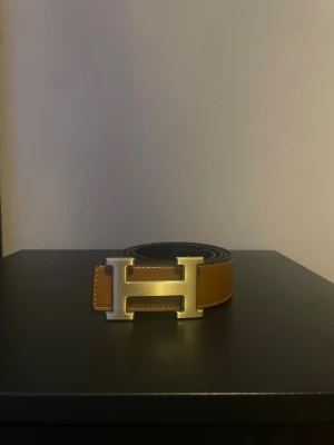  brunt skärp  - Snyggt brunt skärp med ikoniskt H-format spänne i guld. Bältet är gjort i skinn och har kontrastsömmar längs kanterna. Perfekt accessoar för att lyfta vilken outfit som helst.