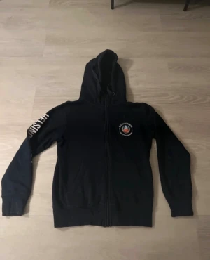 Svart hoodie Helsingborgs IF - Svart hoodie med dragkedja och huva från Helsingborgs IF. Långärmad med vit text 'HELSINGBORGS' längs ena ärmen och klubbmärke på bröstet. Skön och sportig stil, perfekt för dig som gillar fotboll och vill visa ditt lag.saknar ”f” på armen 