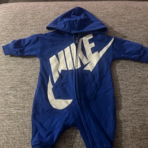 Nike jumper  - Blå hoodie jumpsuit från Nike med stor vit logga framtill och mindre logga baktill. Mjuk bomull, huva, långärmad och dragkedja hela vägen. Praktisk ficka på magen och ribbade muddar vid ärmslut och benslut. Väldigt fint skick då den knappt kommit till användning. 
