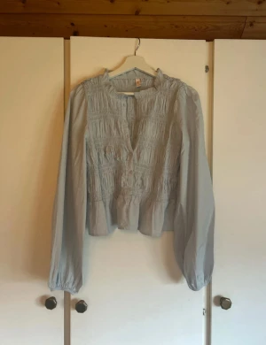 Ljusblå blus med smock från Nelly - Supersöt ljusblå blus från Nelly med smockdetaljer över och volang vid nederkanten. Blusen har långa puffiga ärmar med resår vid handlederna och knappar framtill. Perfekt för dig som gillar romantisk och luftig stil. Den är helt ny och oanvänd tyvärr utan prislapp.
