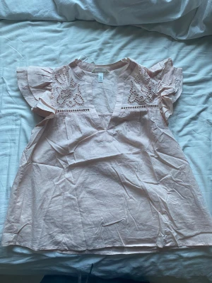 Ljusrosa blus med broderi - Supersöt ljusrosa blus från H&M med v-ringning och broderade detaljer upptill. Blusen har korta volangärmar och är i storlek S. Helt oanvänd!