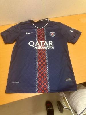 PSG matchtröja Nike blå röd - Paris Saint-Germain fotbollströja från Nike med Qatar Airways tryck på bröstet. Tröjan är marinblå med ett rött och vitt geometriskt mönster längs mitten, PSG-logga och Ligue 1-märke på ärmen. Tillverkad i lätt, tekniskt material med korta ärmar och rund hals.