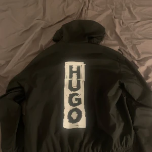 OANVÄND Svart vindjacka från Hugo med tryck - Svart vindjacka från Hugo med stor vit logga på ryggen och mindre logga på bröstet. Jackan har huva med dragsko, dragkedja framtill och elastisk midja. Perfekt för dig som vill ha en stilren och modern look, markerad som M men fungerar som S också.