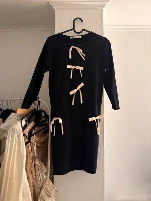 Svart klänning från Zara - Supersöt svart klänning från ZARA med raka linjer och beigea rosetter framtill. Klänningen har trekvartsärm och två fickor framtill.