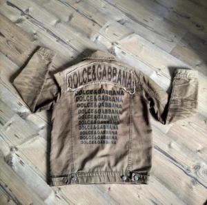 Tvärfet Dolce & Gabbana jacka🔥 - Snygg unik beige jeansjacka från Dolce & Gabbana med tryckt logga i flera rader på ryggen och patchade logodetaljer fram. Klassisk krage, två bröstfickor och knäppning med silvriga knappar. Jackan har en cool, streetig vibe och är tillverkad i bomull. Oerhört niche och rare!