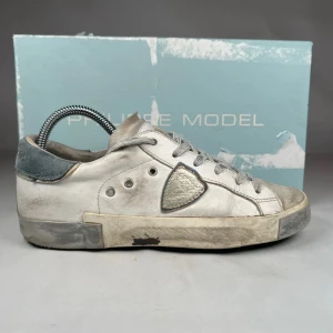 Philippe Model vita sneakers i skinn - Snygga vita sneakers från Philippe Model med grå detaljer och silverfärgad logopatch på sidan. Skorna har rund tå, snörning och platt sula. Tillverkade i skinn med mockadetaljer och coolt sliten look. Perfekta för dig som gillar streetstyle och exklusiva märken. Skoboxen ingår.