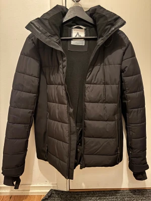 Svart pufferjacka från McKinley - Säljer en svart pufferjacka från McKinley med huva och dragkedja. Jackan har quiltad design, långa ärmar och praktiska fickor med dragkedja. Perfekt för kalla dagar och har en clean, sportig look. Enda defekter är ena tum ”fickan” som går att sy ihop igen!