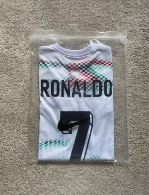 Portugal Ronaldo 7 fotbollströja Puma - Säljer en vit Portugal fotbollströja med Ronaldo 7-tryck på ryggen. Tröjan har korta ärmar, svart krage och detaljer i svart på ärmsluten. Mönstret består av hexagoner i rött och grönt. Puma-logga på bröstet och Portugal-emblem. Materialet är lätt och andas.