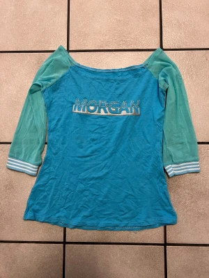 Turkos långärmad Morgan topp storlek Xs/S - Turkos långärmad topp från Morgan med glittrigt logotryck och små pärlor framtill. Ärmarna har ljusare turkos ton och randiga muddar i vitt och turkos. Mjuk och lätt bomullskänsla, perfekt för dig som gillar sportig och snygg stil.