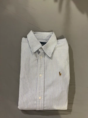 Randig skjorta från Ralph Lauren - Snygg vit och blå randig skjorta från Ralph Lauren med klassisk krage och broderad logga på bröstet. Skjortan har lång ärm, knäppning framtill och är i super slim fit. Perfekt för dig som gillar stilrena och tidlösa plagg.