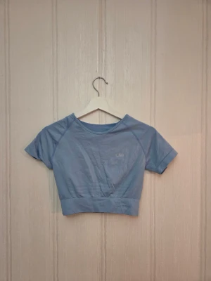 Blå croppad topp från ICANIWILL - Säljer en ljusblå croppad topp från ICANIWILL med korta ärmar och rund halsringning. Diskret logga på bröstet och även uppe vid nacken, bred ribbad kant nedtill för extra support. Stl S, använd fåtal gånger