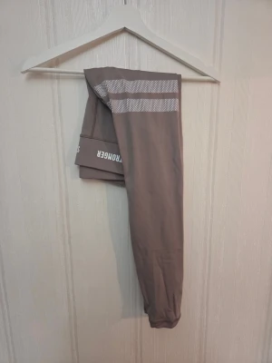Bruna leggings från Stronger - Snygga bruna leggings från Stronger med vita ränder och en tjockare resår upptill. Logga på benet och elastisk midja för extra komfort. Använd fåtall gånger, stl S