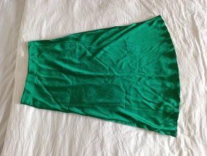 Grön satinkjol från Lindex, M - Snygg grön kjol i satinliknande material från Lindex i storlek M. Kjolen har en lätt glansig yta och är tillverkad i återvunnen polyester. Den har en vågig nederkant som ger en flowig och lätt look.