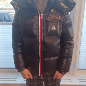 Svart Moncler dunjacka med huva - Säljer en svart, glansig dunjacka från Moncler med stor huva och klassisk logga på bröstet. Jackan har rödvit randig dragkedja, bröstficka med knapp och quiltad design. Perfekt för kalla dagar och riktigt snygg streetstil. Den är i väldigt bra skick då jag har bara använt den några gånger. Säljer den för att den är för liten